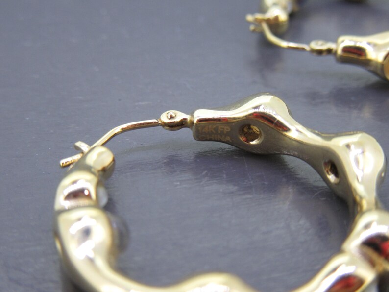 May include: Close-up of gold-colored hoop earrings. The earrings have a unique, textured design with a series of rounded, interconnected shapes. The earrings are marked with "14K FP CHINA". The background is a solid, dark gray color.