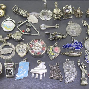 May include: A collection of silver-toned charms, including a telephone, car, guitar, slot machine, and various state and travel-themed pendants. Other charms include a roulette wheel, a heart with a butterfly, and a Statue of Liberty.
