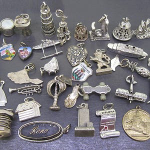May include: A collection of silver-toned charms in various shapes, including a tower, a house, a trumpet, and a Reno oval. Some charms have colorful enamel details. The charms are displayed on a dark gray surface.