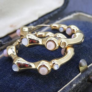 May include: A pair of gold-toned hoop earrings with a series of light pink and white gemstones. The earrings are displayed inside a vintage blue jewelry box, showcasing their design and color contrast.
