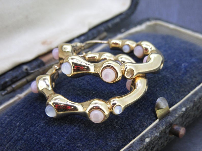May include: A pair of gold-toned hoop earrings with a series of light pink and white gemstones. The earrings are displayed inside a vintage blue jewelry box, showcasing their design and color contrast.