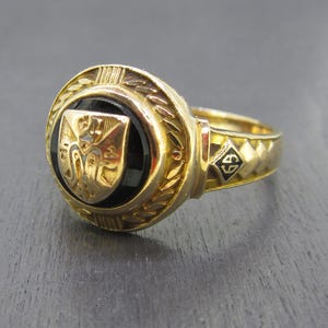 May include: A gold ring featuring a circular black onyx inlay centered with a gold shield emblem. The ring band displays a textured geometric pattern and a small diamond-shaped detail containing the black letters GS on the side of the shank.