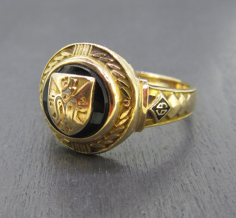 May include: A gold ring featuring a circular black onyx inlay centered with a gold shield emblem. The ring band displays a textured geometric pattern and a small diamond-shaped detail containing the black letters GS on the side of the shank.