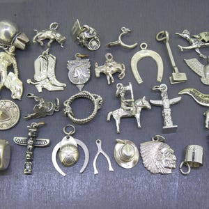 Op de afbeelding: Een verzameling zilverkleurige bedels met Western- en Native American thema's. De bedels omvatten een cowboy, een stier, cowboylaarzen, een hoefijzer, een Native American chief, een totempaal en een cowboyhoed.