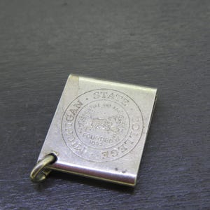 Pu&ograve; includere: Un fermasoldi color argento di forma rettangolare. La clip presenta un emblema circolare con le parole "MICHIGAN STATE COLLEGE" e "FOUNDED 1855". Un piccolo anello &egrave; attaccato in basso.