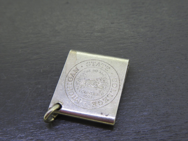 Pu&ograve; includere: Un fermasoldi color argento di forma rettangolare. La clip presenta un emblema circolare con le parole "MICHIGAN STATE COLLEGE" e "FOUNDED 1855". Un piccolo anello &egrave; attaccato in basso.