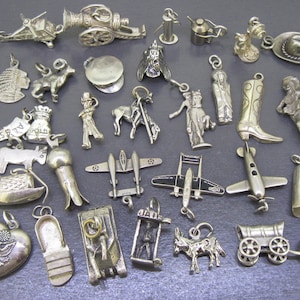 Può includere: Una collezione di ciondoli in metallo color argento di varie forme, tra cui una bandiera americana, uno stivale da cowboy, aerei, un cannone, un cuore con un emblema d'aquila, un carro coperto, un asino, un sombrero e altre figure dettagliate.