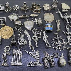 May include: A collection of various silver-toned metal charms featuring diverse shapes including an abacus, a kangaroo, the Eiffel Tower, a ship, a coin, a heart, a beer stein, a piano, a chair, a dancer, and several architectural landmarks.