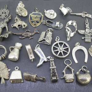 Può includere: Una collezione di ciondoli argentati a tema western e nativo americano. I ciondoli includono uno stivale da cowboy, un ferro di cavallo, un totem, una diligenza, un pozzo, una campana e un roadrunner. I ciondoli sono esposti su una superficie grigio scuro.