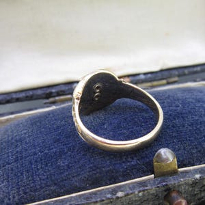 May include: A gold ring featuring a smooth, polished band and a circular head with a dark, recessed center containing two small, vertically aligned metal studs. The ring is positioned inside a dark blue velvet-lined jewelry box.