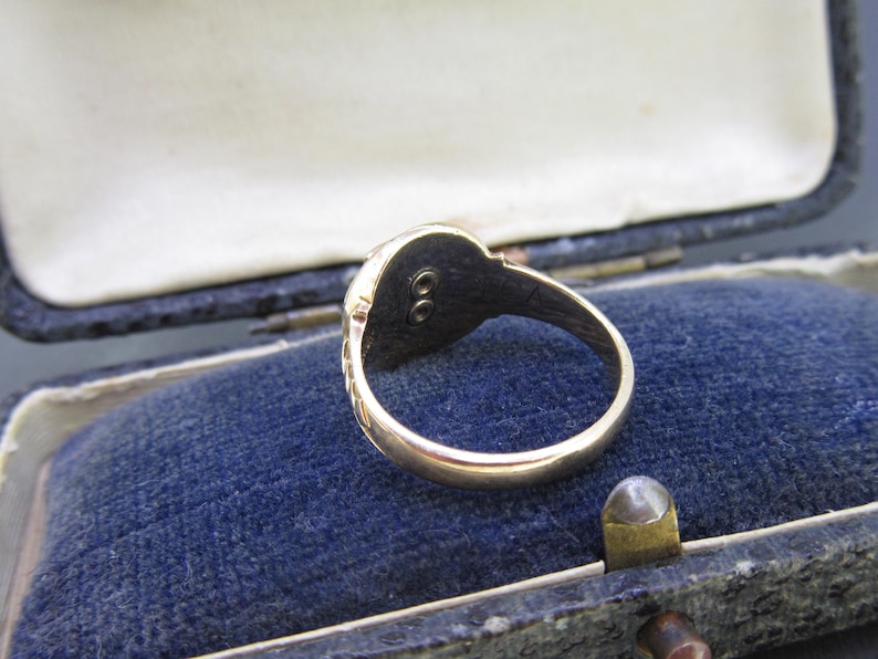May include: A gold ring featuring a smooth, polished band and a circular head with a dark, recessed center containing two small, vertically aligned metal studs. The ring is positioned inside a dark blue velvet-lined jewelry box.