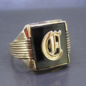 May include: A gold-toned signet ring with a black onyx center stone. The ring features a raised, gold-colored letter "C" in a stylized font. The band has a ribbed design on the sides, adding texture to the classic jewelry piece.