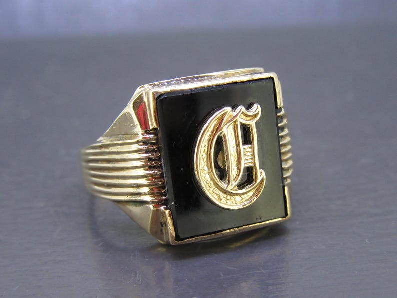 May include: A gold-toned signet ring with a black onyx center stone. The ring features a raised, gold-colored letter "C" in a stylized font. The band has a ribbed design on the sides, adding texture to the classic jewelry piece.
