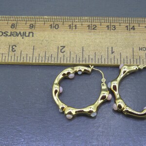 May include: A pair of gold-colored hoop earrings with small, light pink beads. The earrings are next to a wooden ruler, showing measurements in inches. The earrings have a unique, textured design.