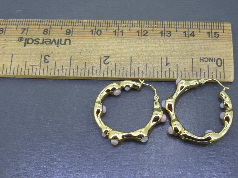 May include: A pair of gold-colored hoop earrings with small, light pink beads. The earrings are next to a wooden ruler, showing measurements in inches. The earrings have a unique, textured design.