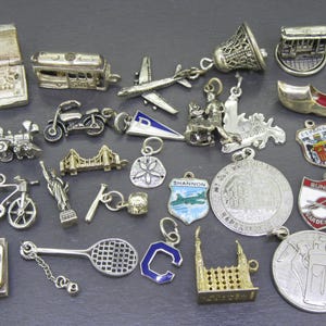 May include: A collection of silver-toned charms, including a train, bicycle, Statue of Liberty, and a tennis racket. Other charms depict a bell, airplane, and a shield with the word "Shannon".