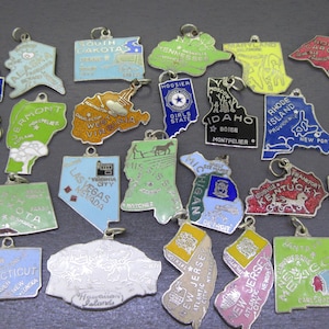 May include: A collection of vintage state souvenir charms in various shapes and colors. Each charm features a state outline, with text indicating the state name and sometimes a city. The charms are made of metal and have a loop for attaching to a chain.