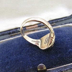 May include: A gold ring with a smooth band and a textured side detail. The interior of the band is engraved with the text JOSTEN 10K and features a small circular symbol with two dots inside. The ring is shown from an angled perspective.