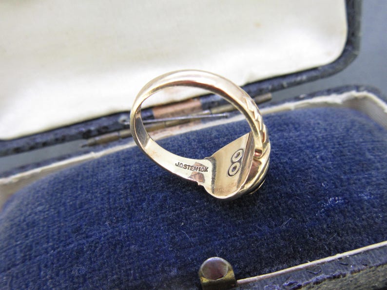 May include: A gold ring with a smooth band and a textured side detail. The interior of the band is engraved with the text JOSTEN 10K and features a small circular symbol with two dots inside. The ring is shown from an angled perspective.