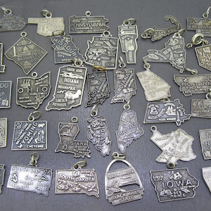 May include: A collection of vintage silver-toned metal state charms, each shaped like a state with engraved details. The charms feature state names, landmarks, and cities. Some have a loop for attaching to a necklace or bracelet.