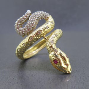 May include: A gold-tone snake ring with clear and red gemstones. The snake is coiled around a band and has a detailed head.