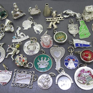 May include: A collection of vintage silver charms, including bells, shields, and pendants with various designs. Some charms feature text like "Happy Anniversary" and "Nebraska". Other charms depict figures, animals, and landmarks.