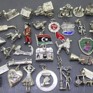May include: A selection of silver charms in diverse shapes and designs. Charms include a guitar, a trolley car, a flag, a heart, a bell, a dog, a figure, and a New Orleans street sign. The charms are displayed on a dark grey surface.