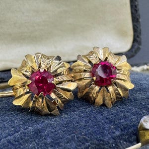 May include: A pair of gold earrings featuring a circular, textured, petal-like design surrounding a central, round, faceted deep red gemstone. The earrings have a polished metallic finish and are set against a dark blue velvet-lined interior.