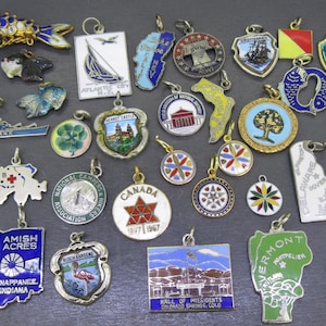 May include: A collection of vintage enamel charms in various shapes and designs. Includes charms with state outlines, landmarks, and nautical themes. The charms are in silver and gold tones with colorful enamel details.