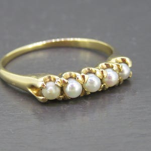 May include: A gold ring featuring a row of five small, round, white pearls set horizontally across the top. The band is smooth and polished, with each pearl held securely in place by individual gold prongs, creating a continuous line design.