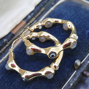 May include: A pair of gold-toned hoop earrings with a unique, textured design. Each earring features multiple clear, rounded stones set along the curved surface. The earrings are displayed in a blue velvet jewelry box.