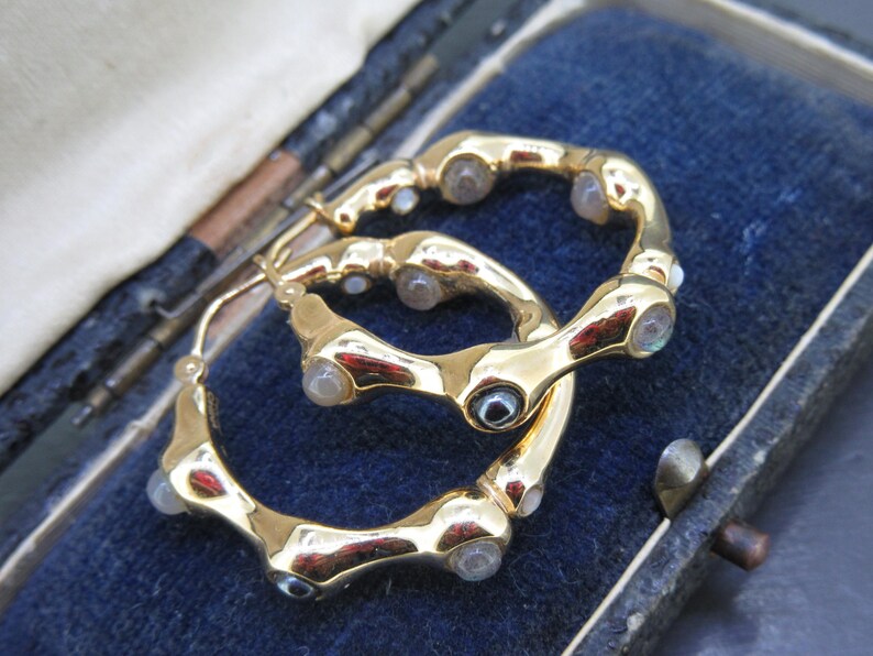 May include: A pair of gold-toned hoop earrings with a unique, textured design. Each earring features multiple clear, rounded stones set along the curved surface. The earrings are displayed in a blue velvet jewelry box.