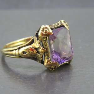 May include: A gold ring featuring a rectangular, faceted purple gemstone set in a decorative metal mounting. The band and setting display intricate, raised floral and scrollwork details that frame the central stone on both sides.