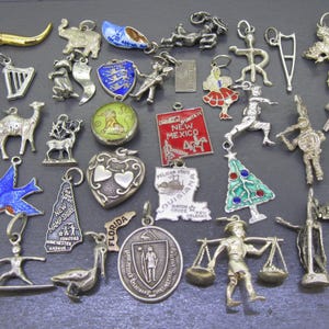 May include: A collection of various metal charms including a gold-toned horn, a blue and white clog, a silver camel, a red New Mexico square, a heart-shaped charm, a Louisiana state shape, a Christmas tree, and a person carrying scales.