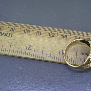 May include: A gold-colored ring with a black rectangular stone is shown next to a wooden ruler. The ring has a detailed design on the side and is approximately 0.75 inches in diameter. The ruler is marked in inches.
