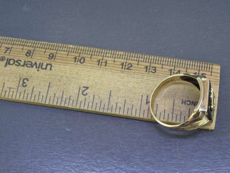 May include: A gold-colored ring with a black rectangular stone is shown next to a wooden ruler. The ring has a detailed design on the side and is approximately 0.75 inches in diameter. The ruler is marked in inches.