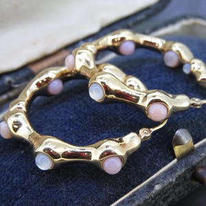 May include: A pair of gold-toned hoop earrings with a unique design. Each earring features a series of rounded segments, interspersed with small, round, pink and white gemstones. The earrings are displayed on a dark blue surface.