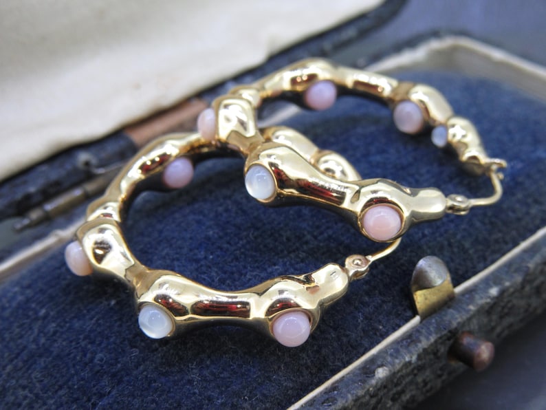 May include: A pair of gold-toned hoop earrings with a unique design. Each earring features a series of rounded segments, interspersed with small, round, pink and white gemstones. The earrings are displayed on a dark blue surface.