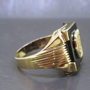 May include: A gold-colored ring with a black rectangular stone and a circular emblem. The ring has a ribbed design on the band and a detailed setting around the black stone. The emblem appears to be gold-colored.