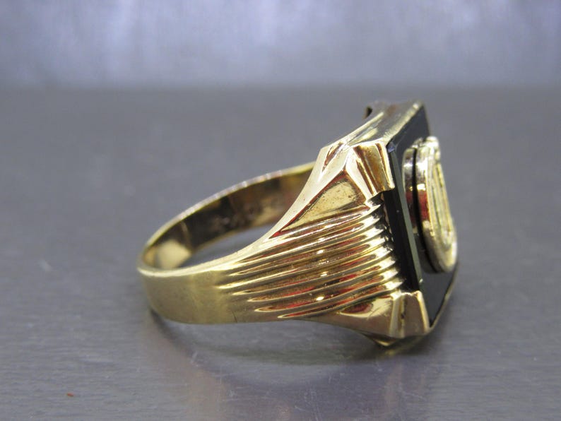 May include: A gold-colored ring with a black rectangular stone and a circular emblem. The ring has a ribbed design on the band and a detailed setting around the black stone. The emblem appears to be gold-colored.