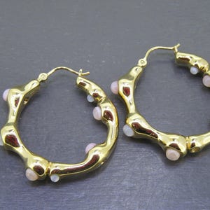 May include: A pair of gold-colored hoop earrings. The earrings have an irregular, textured design with small, light pink and white accents. The hoops are approximately 1.5 inches in diameter.