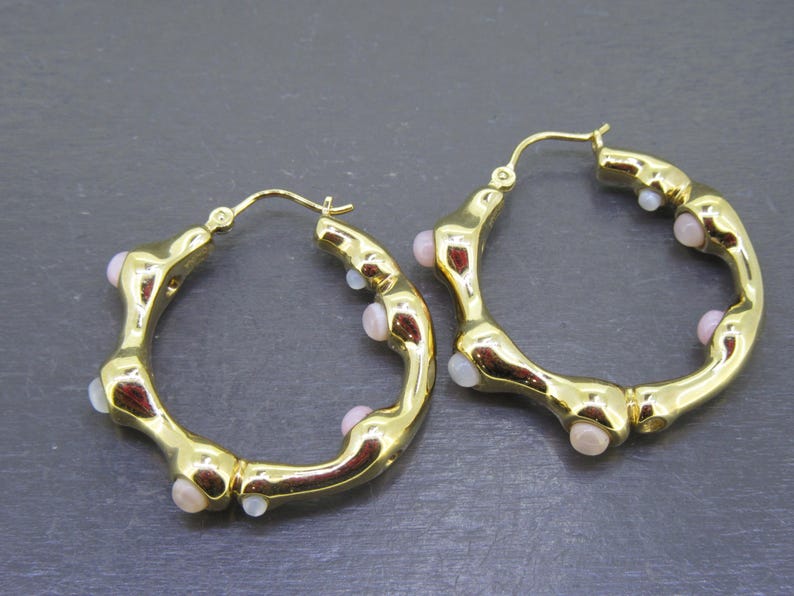May include: A pair of gold-colored hoop earrings. The earrings have an irregular, textured design with small, light pink and white accents. The hoops are approximately 1.5 inches in diameter.