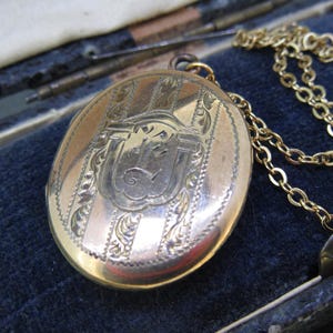 May include: A gold-toned oval locket necklace with an engraved design. The locket features a shield-like emblem and vertical stripes. The chain is a delicate gold color, and the locket rests on a dark blue velvet surface.