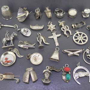 May include: A selection of small, silver-coloured charms and pendants. The collection includes a thimble, a bird, a guitar, a cowboy on a horse, a wheel, a horseshoe, and other Western-themed and novelty designs. The charms are detailed and have a vintage appearance.