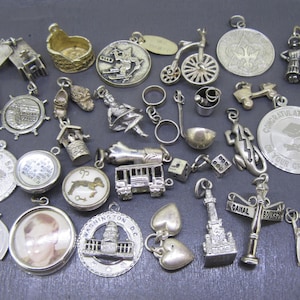 Peut inclure: Une collection de breloques en argent vintage de différentes formes et tailles. Comprend des breloques d'un vélo, d'un puits, d'une tour Eiffel miniature, d'un médaillon de remise de diplôme et d'un charme en forme de cœur. Les breloques sont exposées sur une surface sombre.
