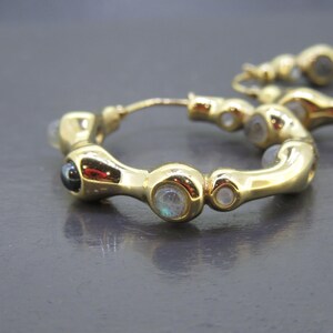 May include: Gold-toned hoop earrings with a series of small, round gemstones. The earrings have a unique, curved design, with a mix of clear and dark gemstones. The earrings are displayed on a dark gray surface.