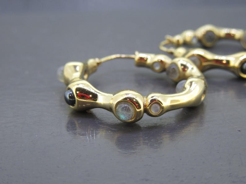 May include: Gold-toned hoop earrings with a series of small, round gemstones. The earrings have a unique, curved design, with a mix of clear and dark gemstones. The earrings are displayed on a dark gray surface.