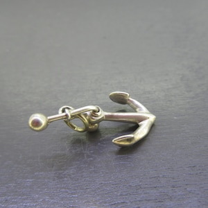 Pu&ograve; includere: Un ciondolo a forma di ancora color argento con una piccola sfera attaccata da una catena. L'ancora presenta un design classico con un anello e due bracci curvi. Il ciondolo &egrave; probabilmente in metallo ed &egrave; posto su uno sfondo scuro.