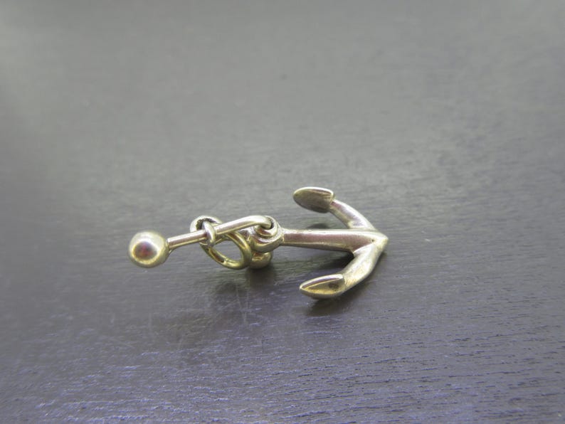 Pu&ograve; includere: Un ciondolo a forma di ancora color argento con una piccola sfera attaccata da una catena. L'ancora presenta un design classico con un anello e due bracci curvi. Il ciondolo &egrave; probabilmente in metallo ed &egrave; posto su uno sfondo scuro.