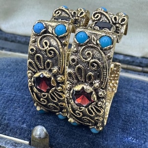 May include: A pair of gold-toned hoop earrings featuring an intricate scrollwork pattern. Each earring is set with a central faceted deep red garnet gemstone and accented with multiple small, round, bright turquoise cabochon stones.